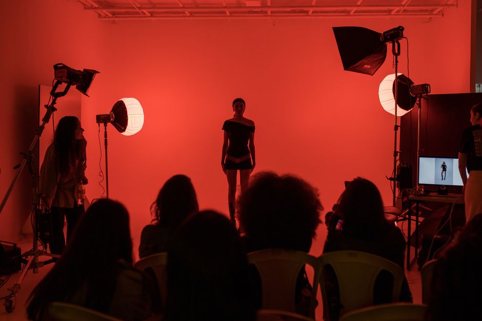 A fashion model poses in a studio with dramatic red lighting while an audience observes the scene.