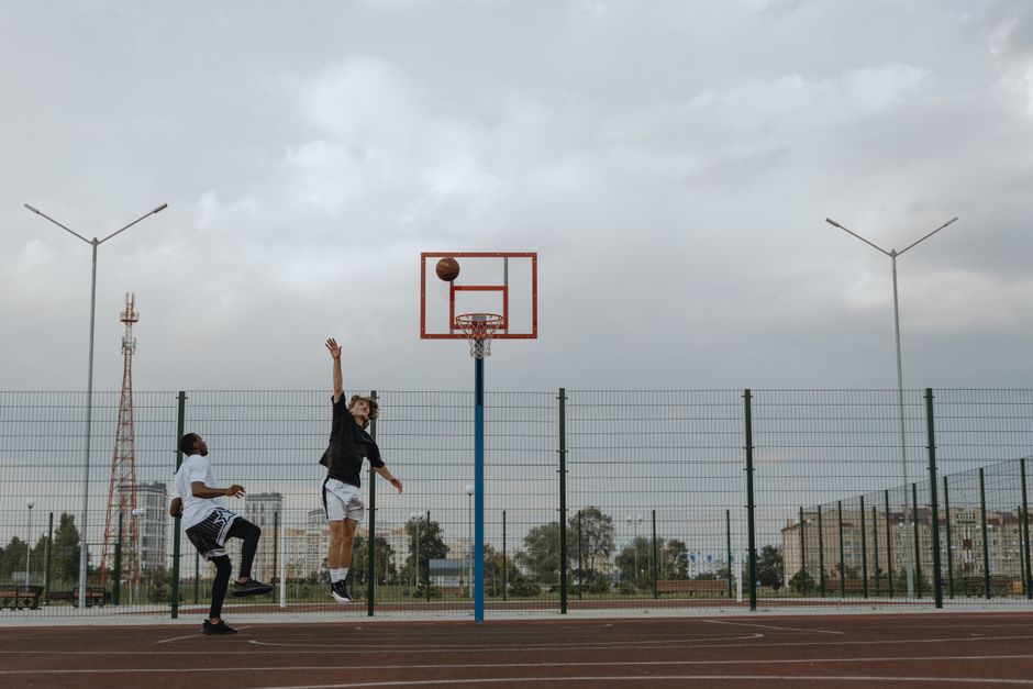 Two men playing basketball outdoors, jumping for a basket on a cloudy day.