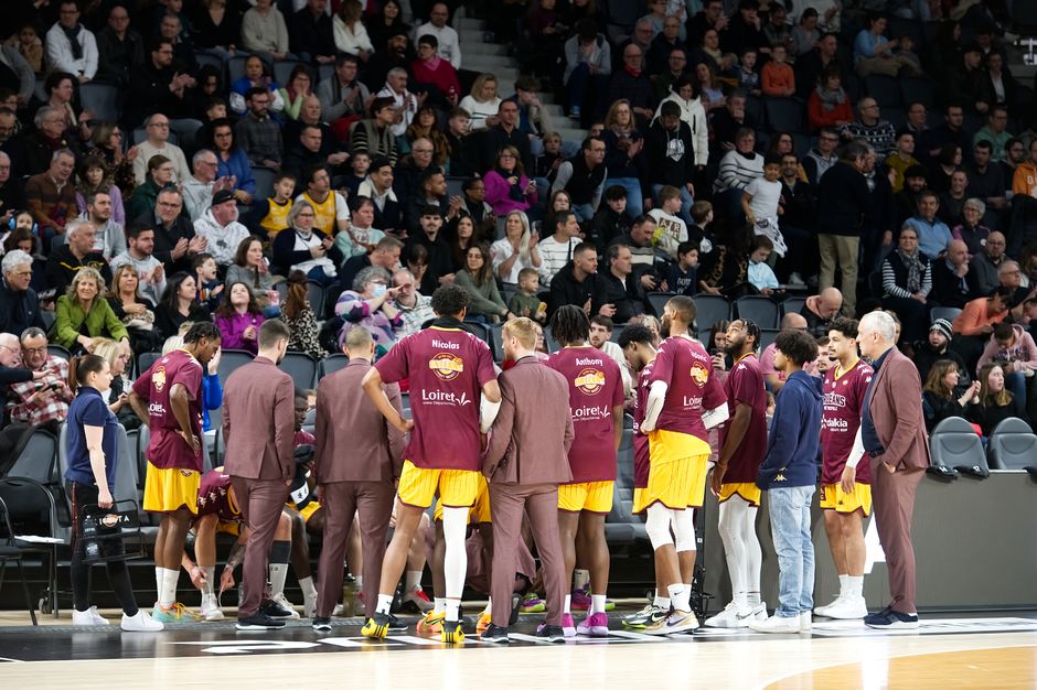 Basketball team huddles on court during a game timeout with audience in background.
