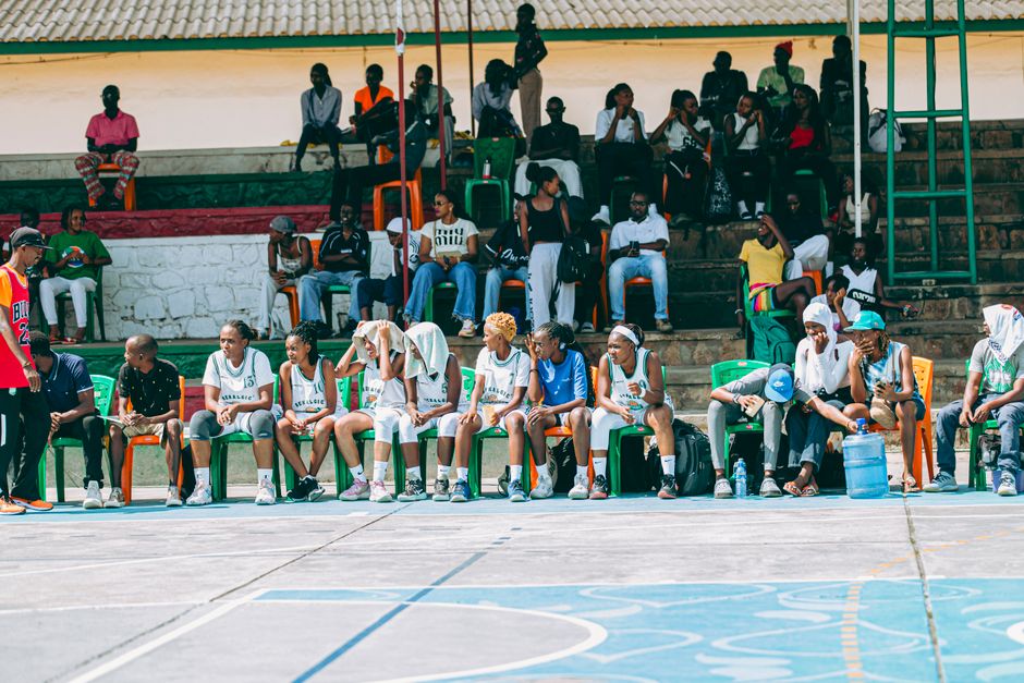 Team sitting on sidelines at outdoor basketball game with spectators in background.