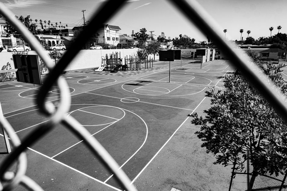 Black and white image of an outdoor basketball court viewed through a chain-link fence.