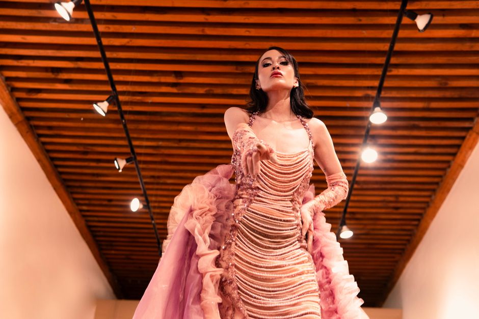 A confident model showcases a stylish pink gown on the runway with dramatic wooden ceiling lighting.